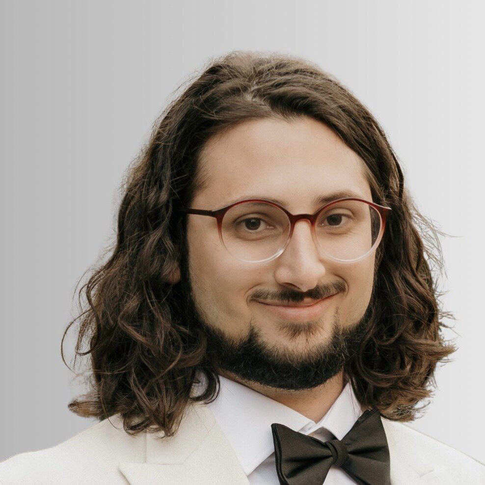 Man with glasses and long hair wearing a white tuxedo and bow tie.